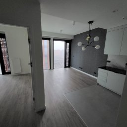 Mateusz Marcinkiewicz - Nowoczesne wnętrze mieszkania z szarą tapetą w geometryczne wzory, drewnianą podłogą i minimalistyczną zabudową kuchenną w bieli. Widok z korytarza.