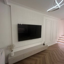 Mateusz Marcinkiewicz - Eleganckie wykończenie ściany z panelem dekoracyjnym i telewizorem. Pod spodem szafka RTV z subtelnymi, złotymi uchwytami. Parkiet w jodełkę i schody z podświetleniem LED.