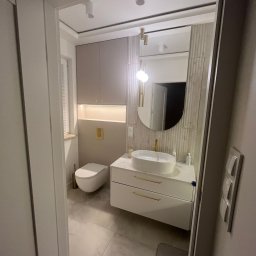 Mateusz Marcinkiewicz - Nowoczesna łazienka z owalnym lustrem, białą umywalką na szafce z złotymi uchwytami i podwieszaną toaletą. Stonowana kolorystyka i oświetlenie punktowe.