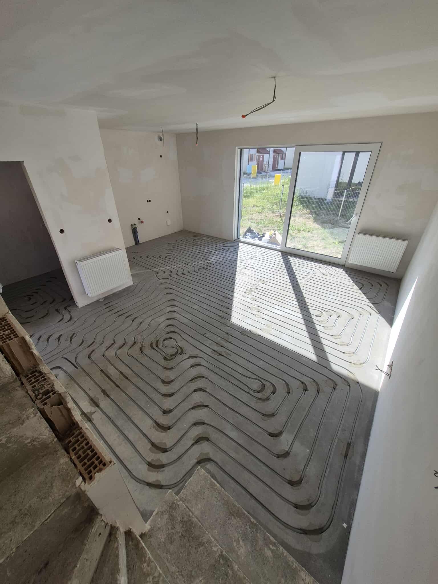 Szare rury ogrzewania podłogowego ułożone w zygzak na wylewce w jasnym pomieszczeniu z oknem balkonowym i grzejnikiem. Widoczne fragmenty schodów i ściany w trakcie wykończenia.