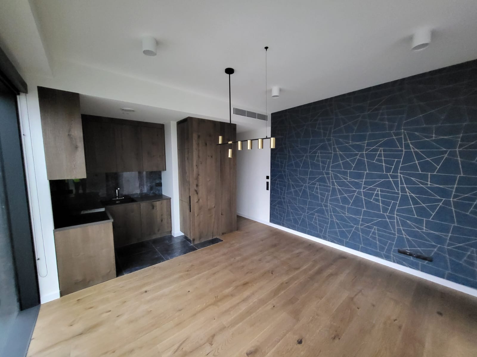 Nowoczesne wnętrze po remoncie: drewniana podłoga, zabudowa kuchenna z ciemnego drewna i ściana z geometrycznym wzorem w odcieniach granatu i złota.