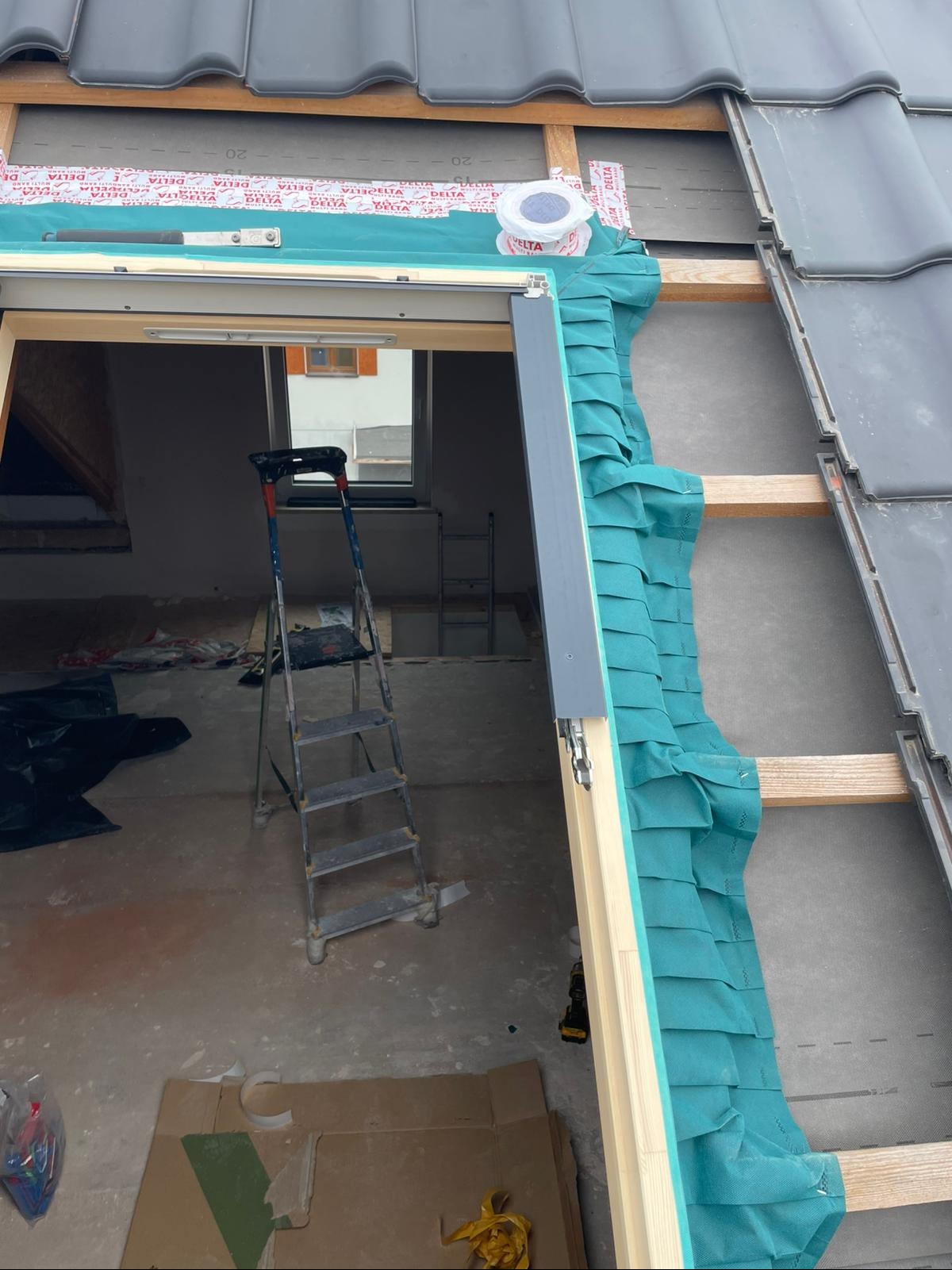 Montaż okna dachowego z uszczelnieniem i folią paroizolacyjną. Widoczna konstrukcja dachu i drabina w surowym wnętrzu poddasza.