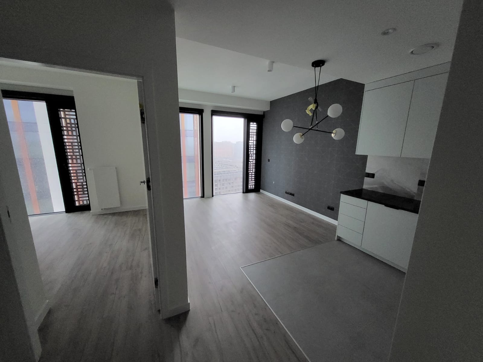 Nowoczesne wnętrze mieszkania z szarą tapetą w geometryczne wzory, drewnianą podłogą i minimalistyczną zabudową kuchenną w bieli. Widok z korytarza.