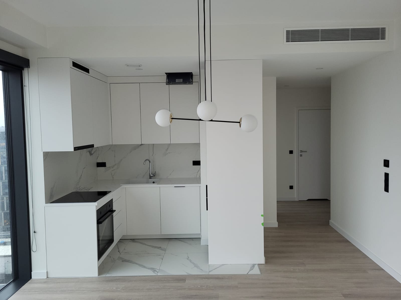Nowoczesna, minimalistyczna kuchnia z białymi szafkami, marmurowym blatem i podłogą. Czarna lampa wisząca z trzema białymi kulami. Widok na korytarz.