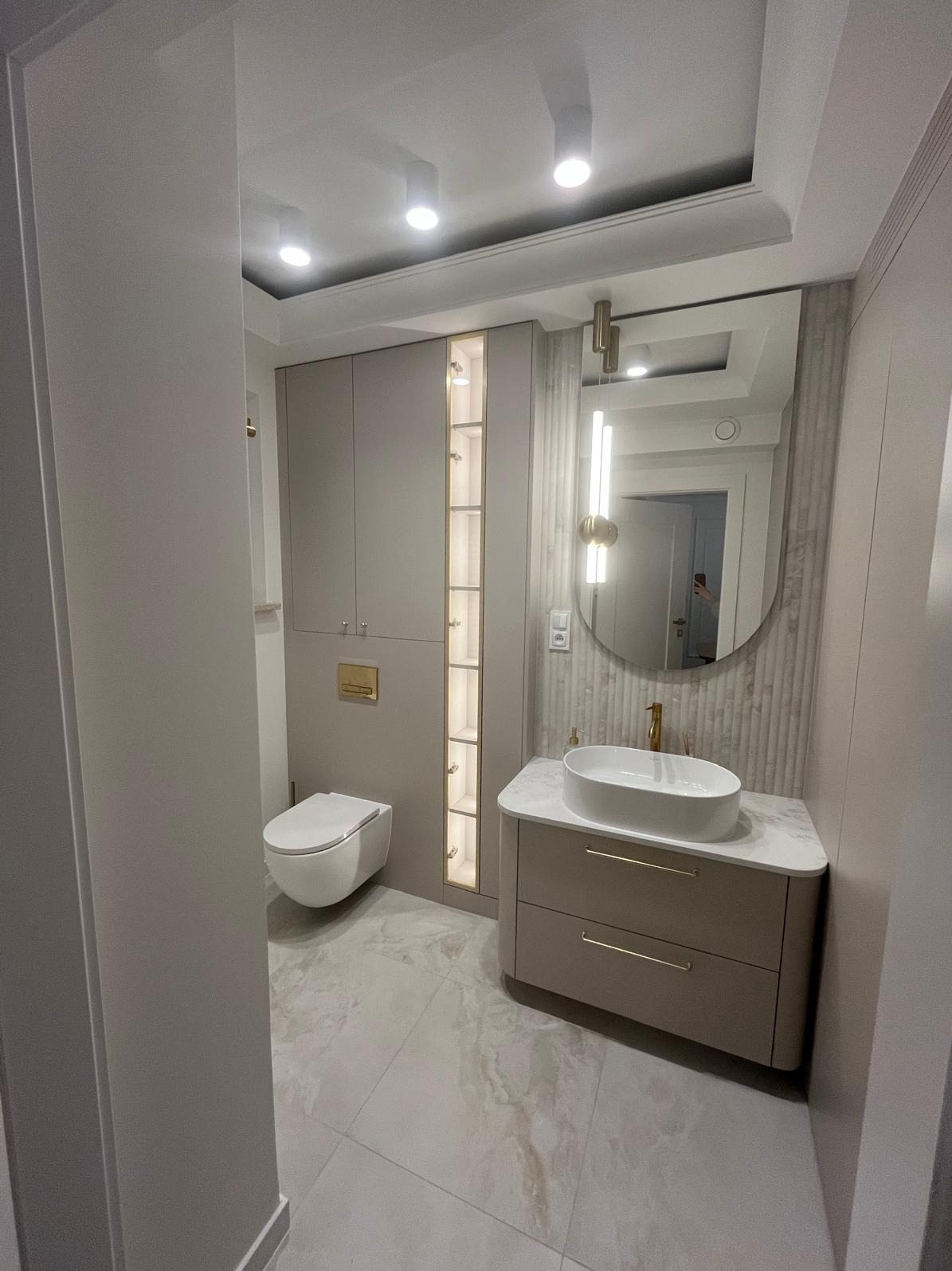 Elegancka łazienka w odcieniach beżu z owalną umywalką na szafce, podświetlaną szafką w zabudowie i dużym lustrem. Minimalistyczny design, płytki imitujące marmur.