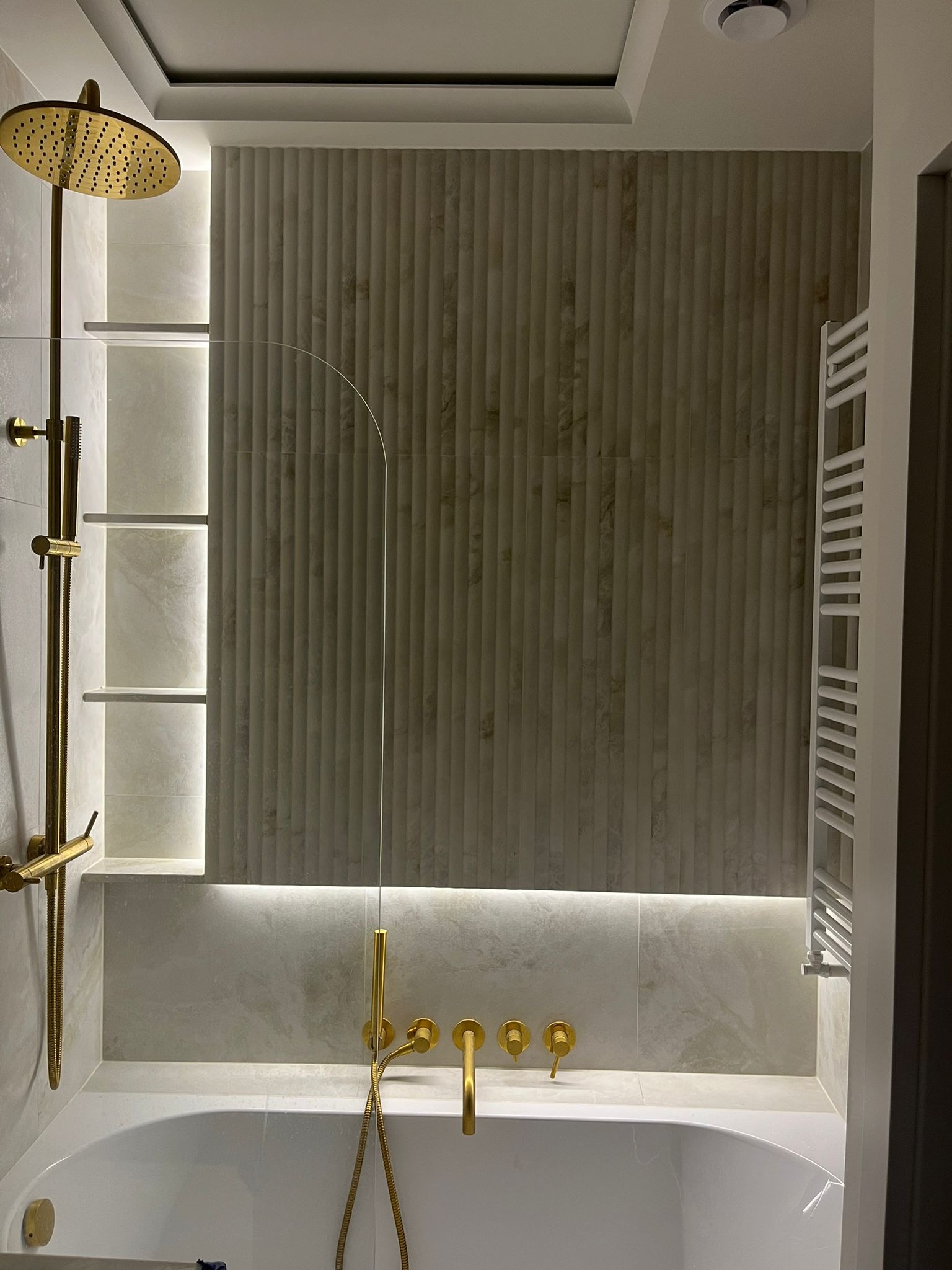 Nowoczesna łazienka w Warszawie: złota armatura, podświetlane półki i dekoracyjne płytki ścienne tworzą elegancki klimat. Biały grzejnik drabinkowy.