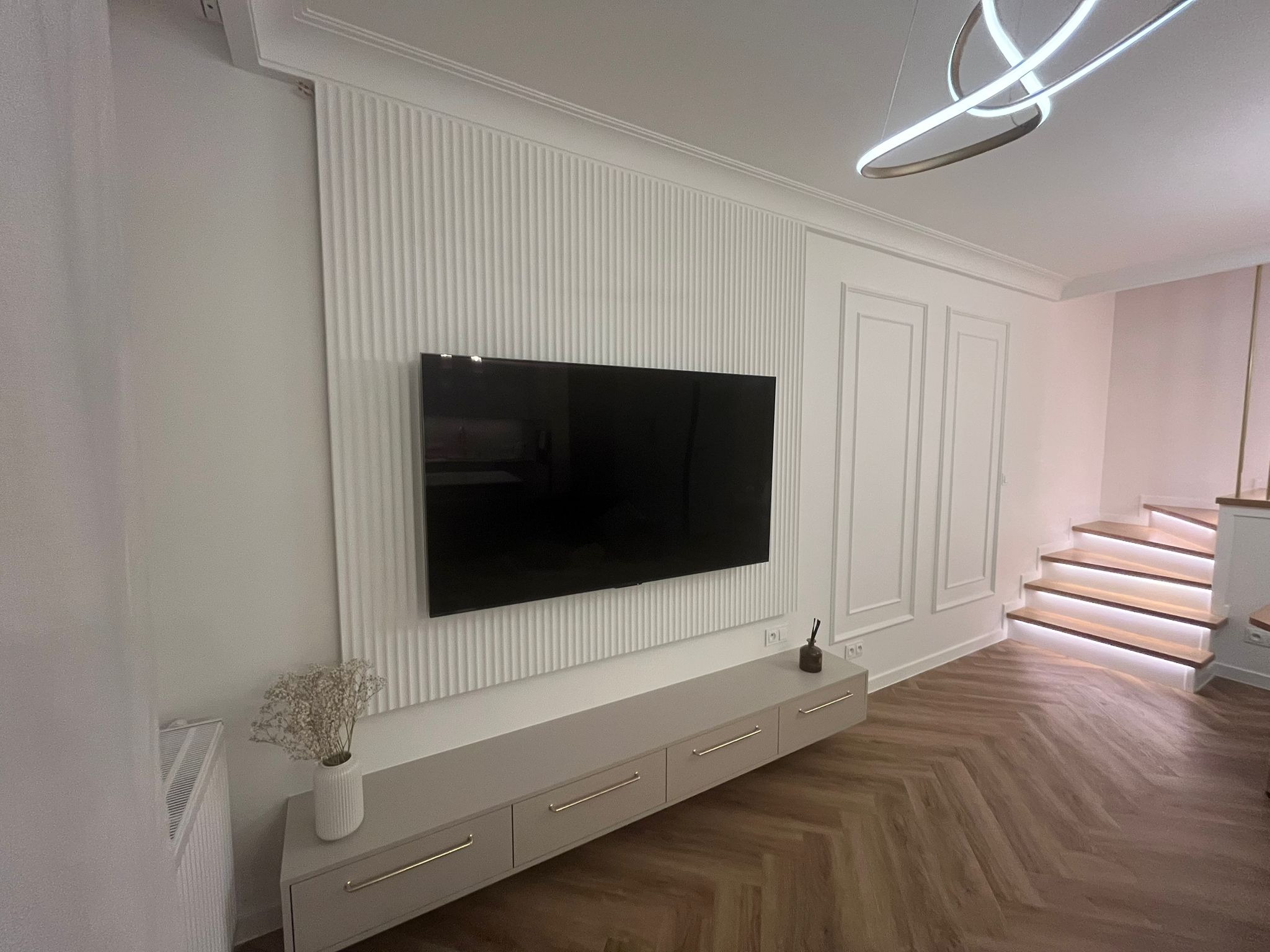 Eleganckie wykończenie ściany z panelem dekoracyjnym i telewizorem. Pod spodem szafka RTV z subtelnymi, złotymi uchwytami. Parkiet w jodełkę i schody z podświetleniem LED.