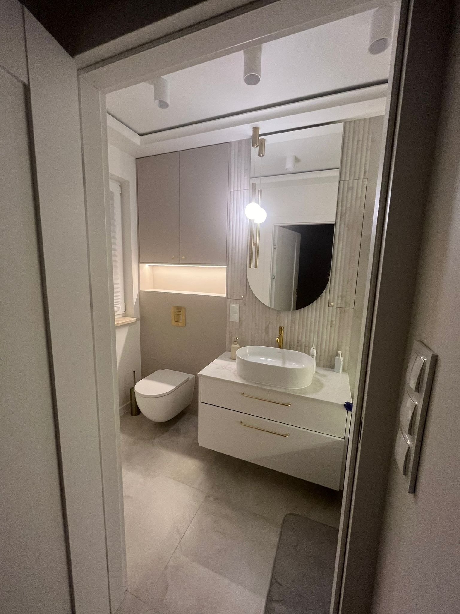 Nowoczesna łazienka z owalnym lustrem, białą umywalką na szafce z złotymi uchwytami i podwieszaną toaletą. Stonowana kolorystyka i oświetlenie punktowe.