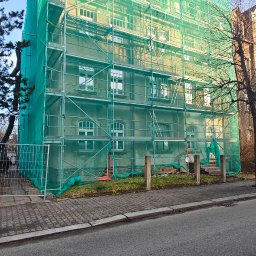 Optimal Tech Rusztowania - Budynek w rusztowaniach, osłonięty zieloną siatką ochronną. Widok z poziomu ulicy, fragment chodnika i drzewa. Prace remontowe lub budowlane w toku.