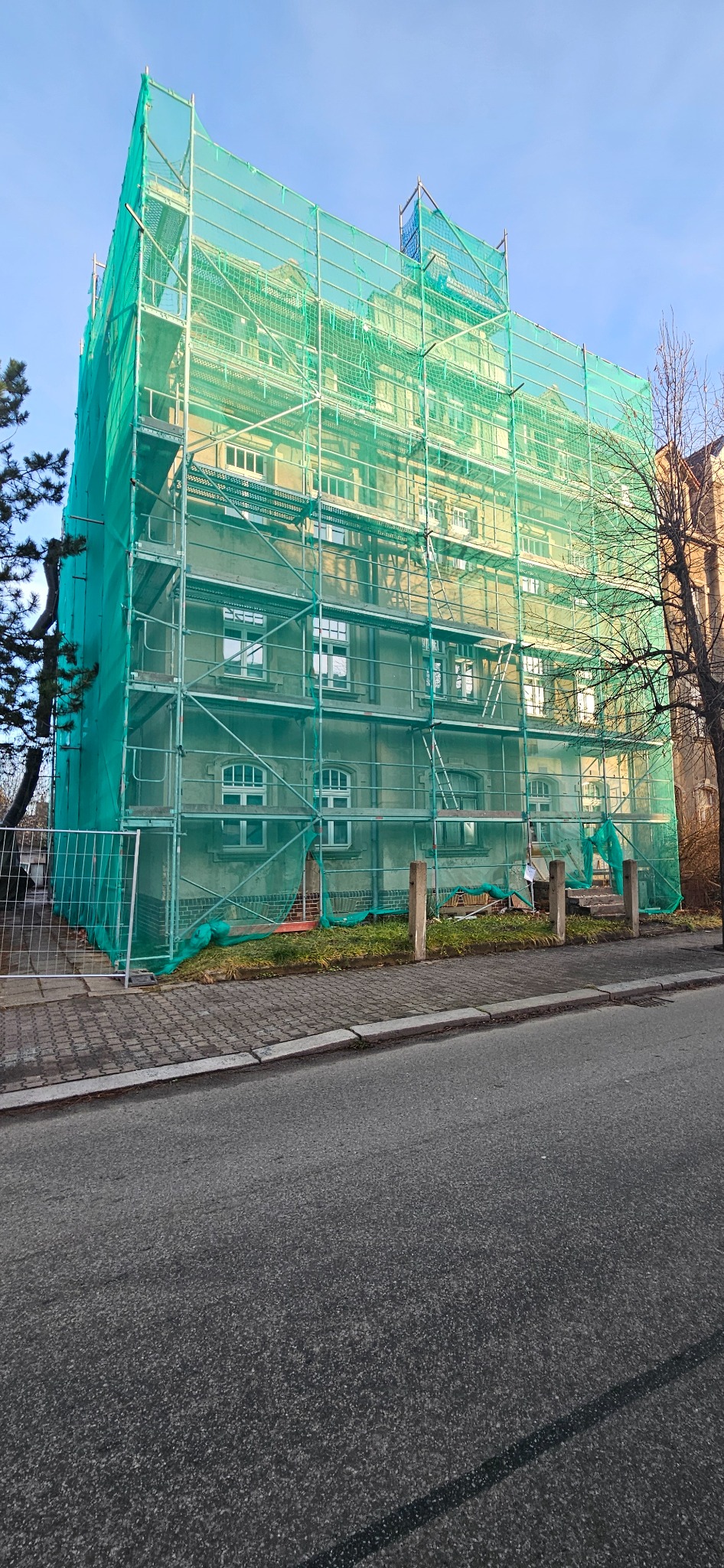 Budynek w rusztowaniach, osłonięty zieloną siatką ochronną. Widok z poziomu ulicy, fragment chodnika i drzewa. Prace remontowe lub budowlane w toku.