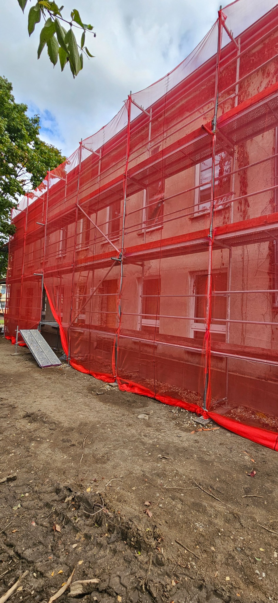 Budynek w rusztowaniach z czerwoną siatką ochronną, widok z dołu. Na pierwszym planie ziemia, w tle zieleń drzew i fragment nieba.