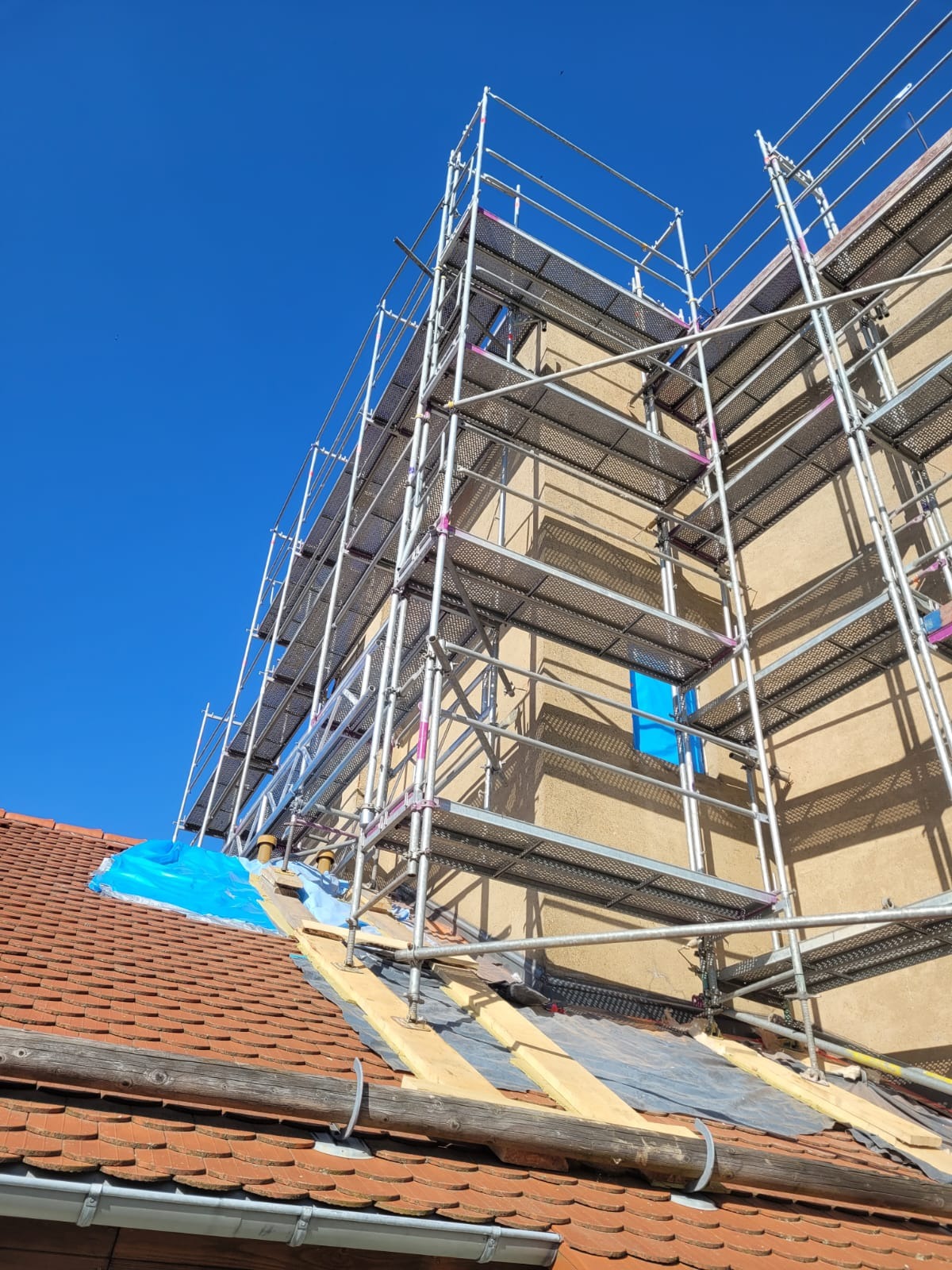 Metalowe rusztowanie na tle błękitnego nieba, częściowo zasłaniające elewację budynku z oknem zabezpieczonym folią i fragment dachu z czerwoną dachówką.