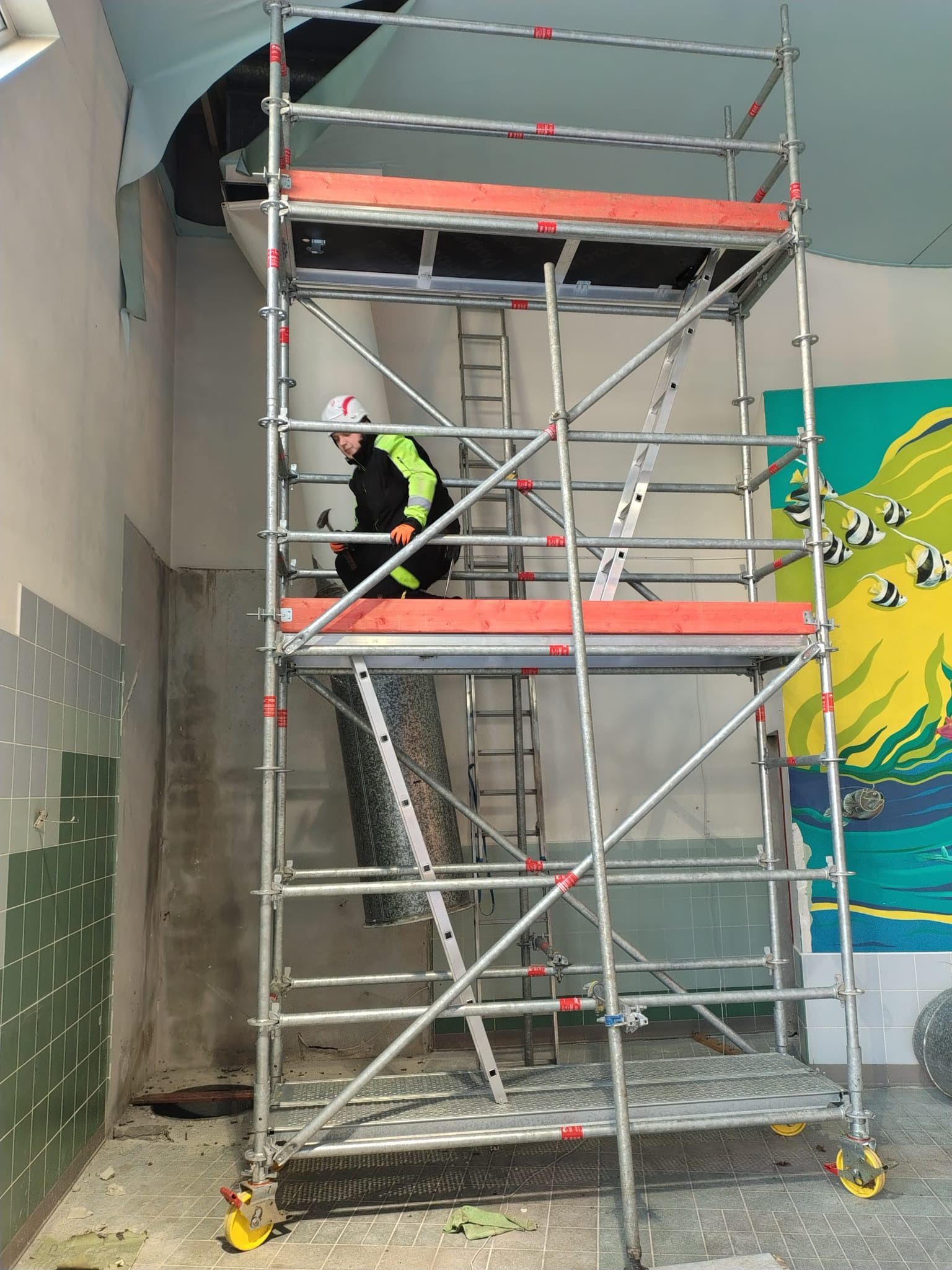 Pracownik w kasku na rusztowaniu wieżowym na kółkach, z młotkiem w dłoni, podczas prac remontowych w pomieszczeniu z muralem. Widoczne płytki i fragment sufitu.