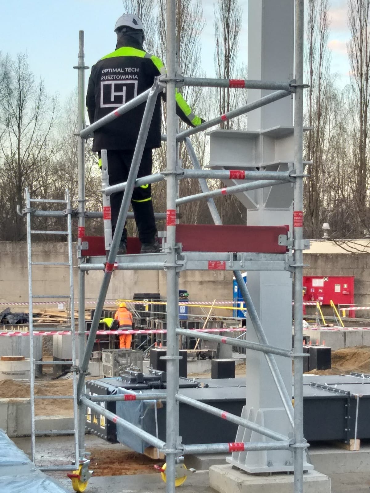 Pracownik w hełmie na rusztowaniu modułowym, w tle konstrukcja stalowa i teren budowy. Widoczny logotyp firmy na kurtce. Ujęcie z dołu.