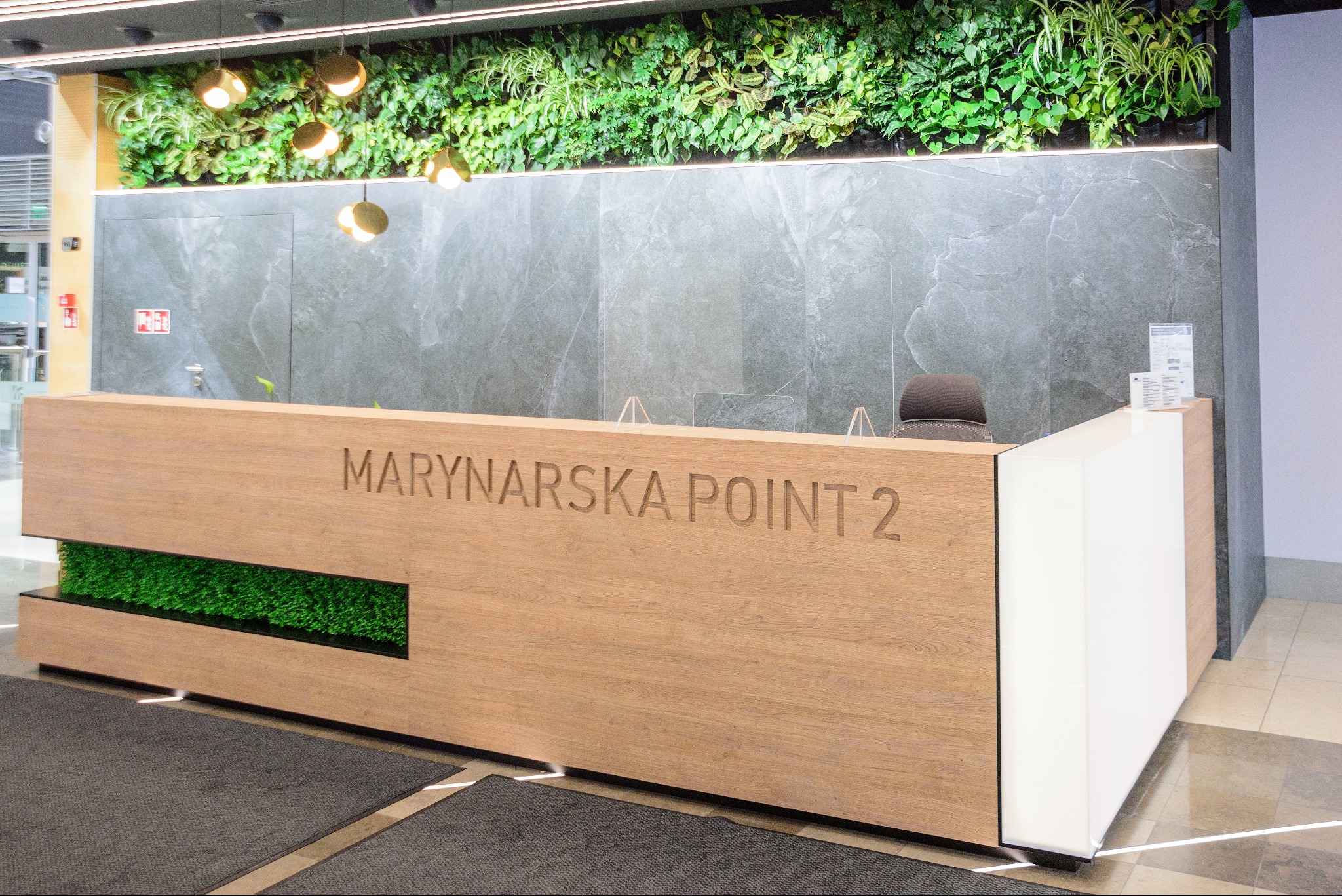 Recepcja biurowa Marynarska Point 2 z drewnianym frontem i zielonym panelem, na tle ściany z roślinami i marmuru. Nowoczesny design, oświetlenie punktowe.
