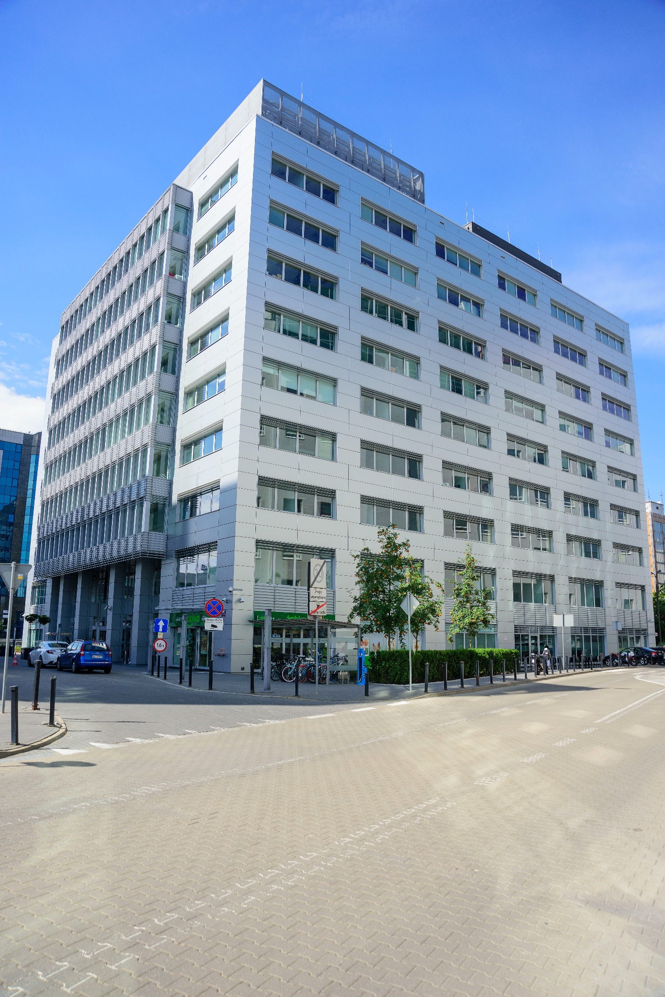 Nowoczesny, srebrny biurowiec z licznymi oknami, widziany z poziomu ulicy w słoneczny dzień. Na pierwszym planie chodnik z kostki brukowej i znaki drogowe.