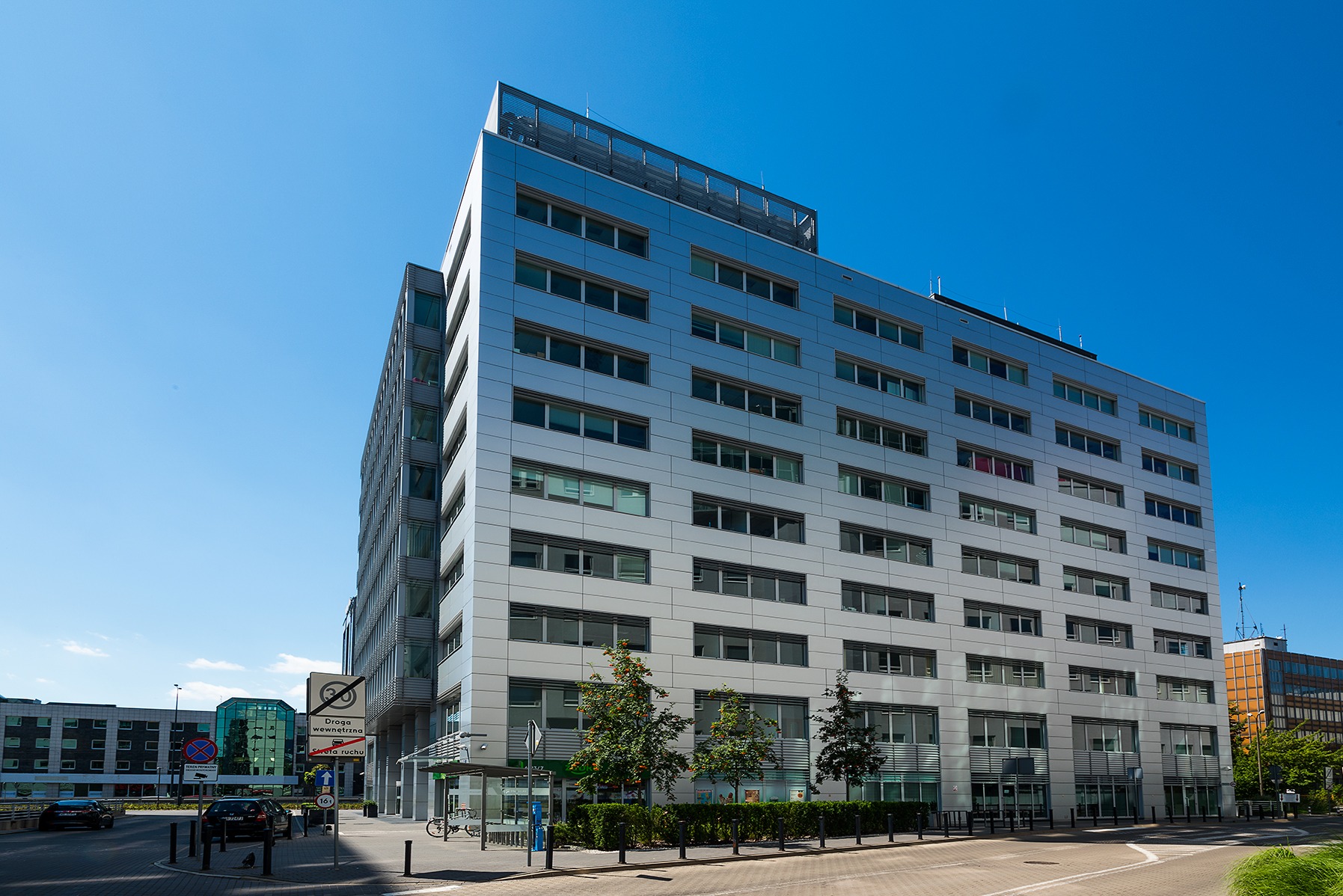 Nowoczesny, srebrny biurowiec z licznymi oknami, widziany z poziomu ulicy w słoneczny dzień. Widoczne drzewa i elementy infrastruktury miejskiej.