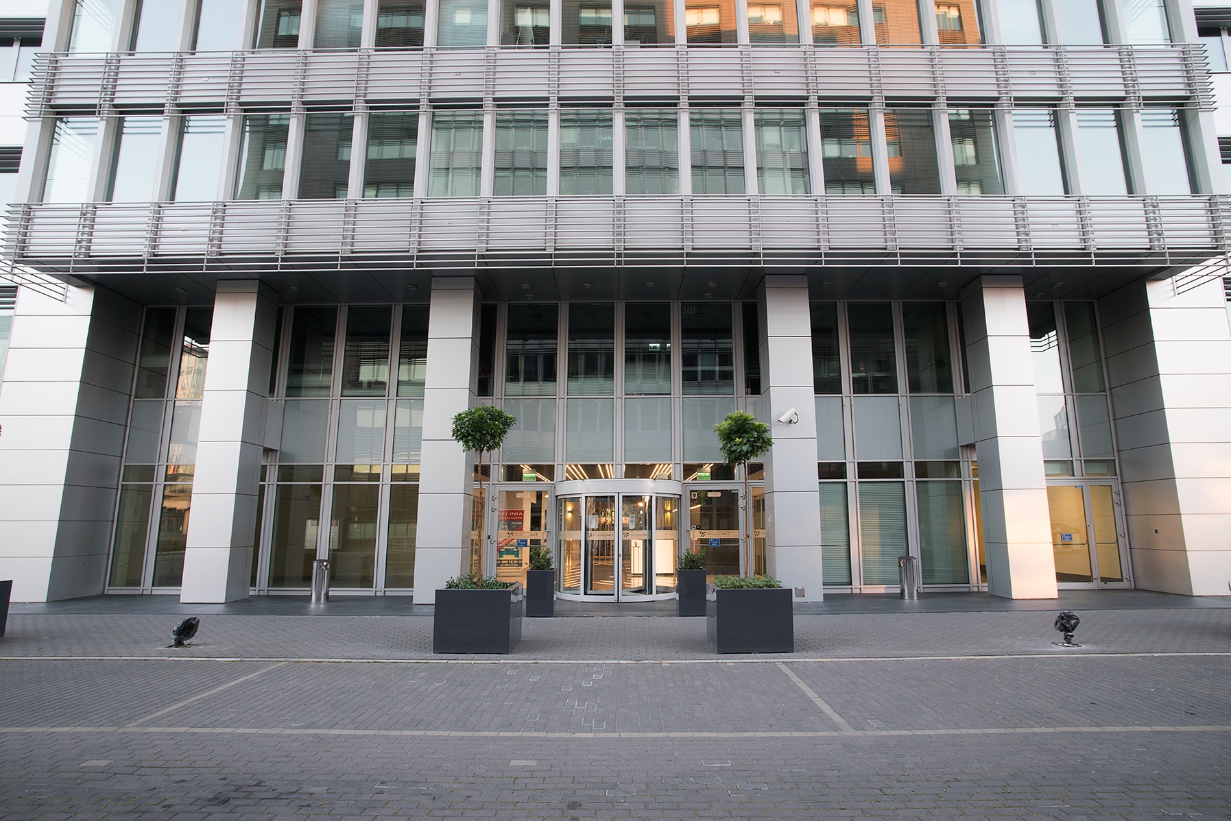 Nowoczesny budynek biurowy z obrotowymi drzwiami wejściowymi, symetrycznie ustawionymi donicami z drzewkami i szarą kostką brukową przed wejściem.
