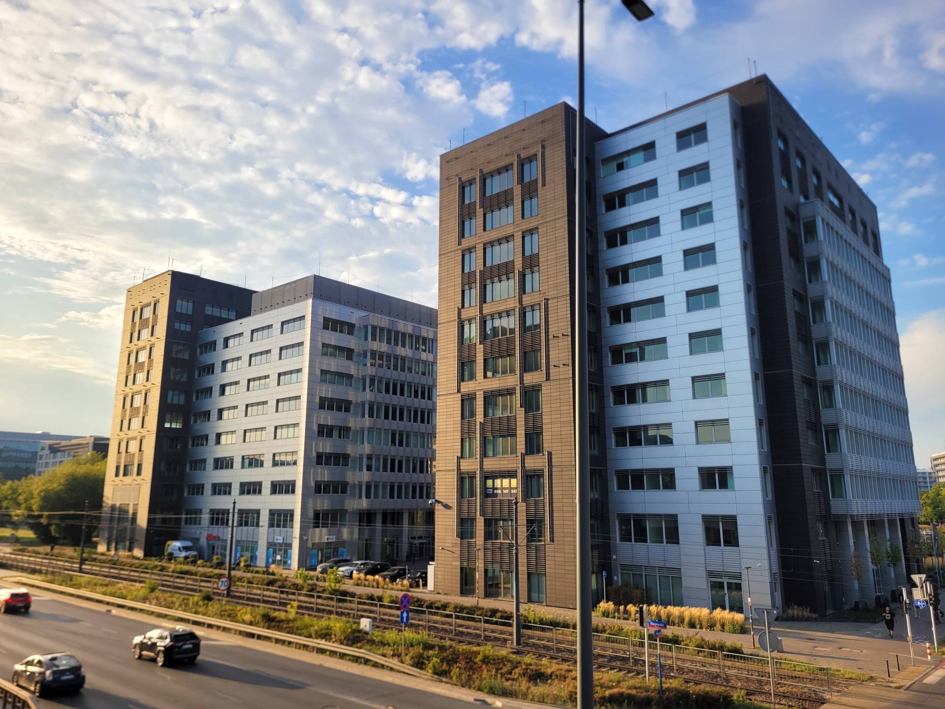 Dwa nowoczesne biurowce z elewacją w odcieniach szarości i brązu, widoczne z perspektywy ulicy z ruchem samochodowym i torami kolejowymi. Błękitne niebo z chmurami w tle.
