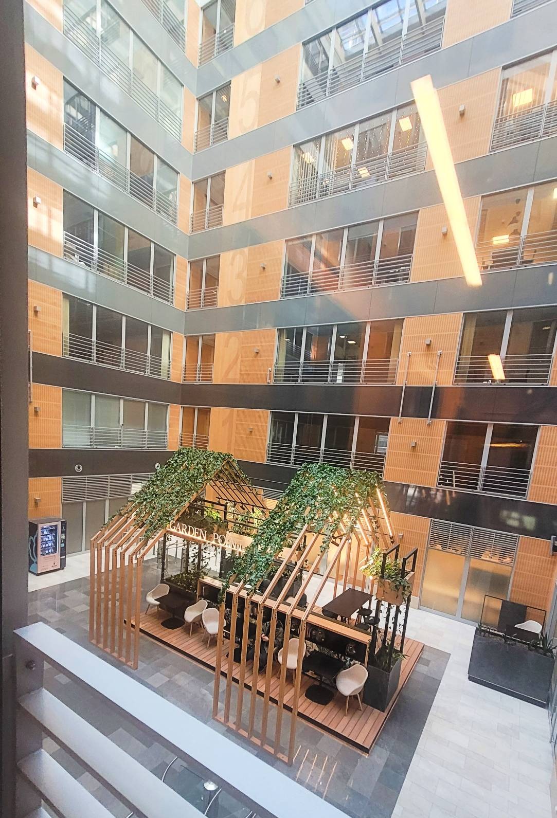 Wnętrze biurowca z przeszklonym atrium, w którym znajduje się drewniana konstrukcja z roślinami, tworząca strefę relaksu z krzesłami i stolikami.