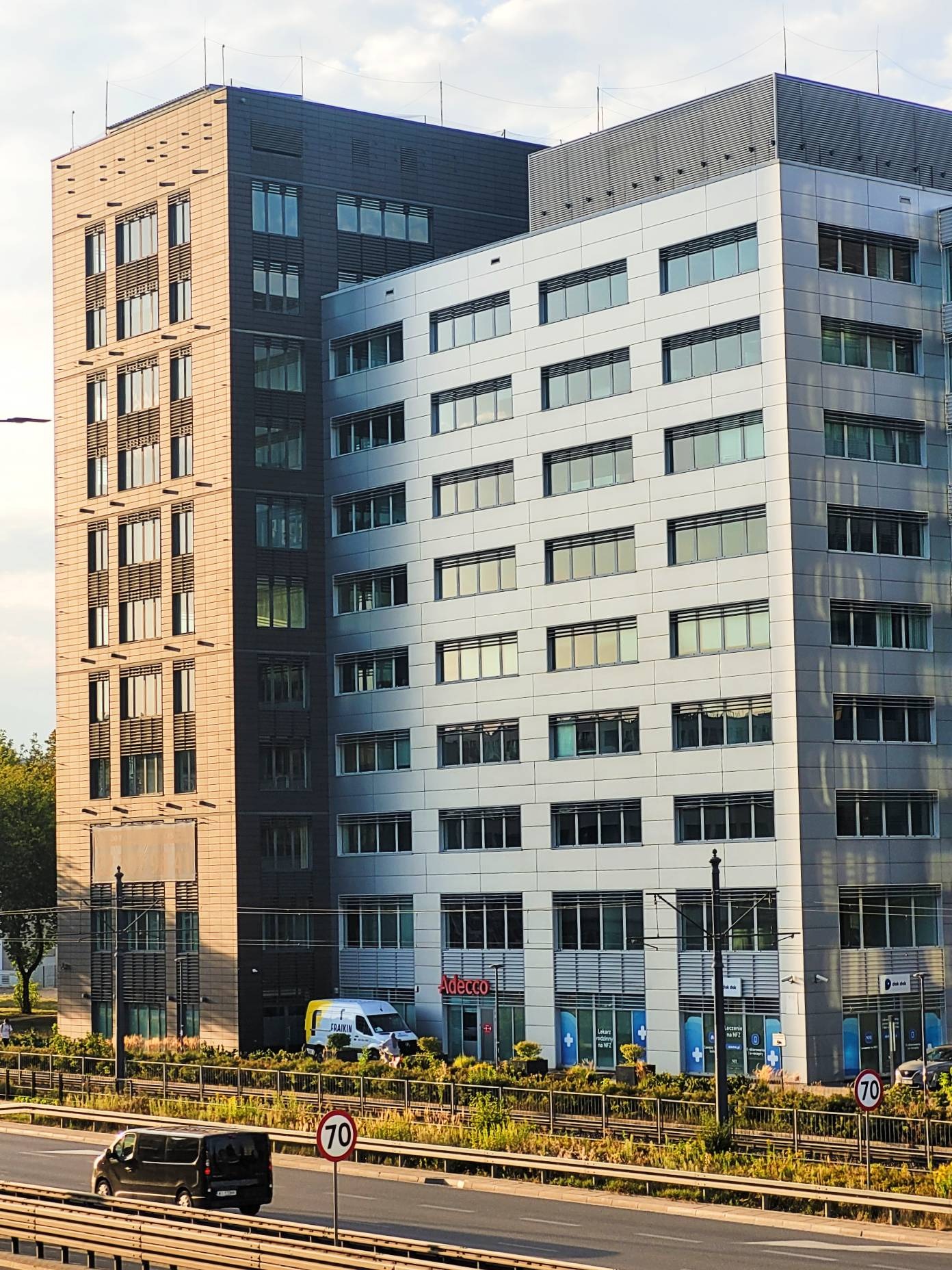 Nowoczesny biurowiec z przeszkloną fasadą i logo Adecco, widoczny z perspektywy ulicy. Uwagę zwraca kontrast między jasną i ciemną częścią budynku.