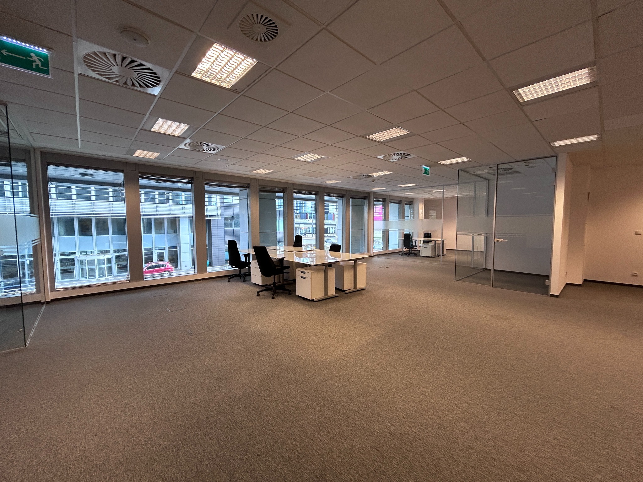 Przestronne biuro z dużymi oknami, widok na zewnątrz. Wnętrze z biurkami i krzesłami, oświetlenie sufitowe. Wykładzina dywanowa na podłodze. Szklane ściany działowe.