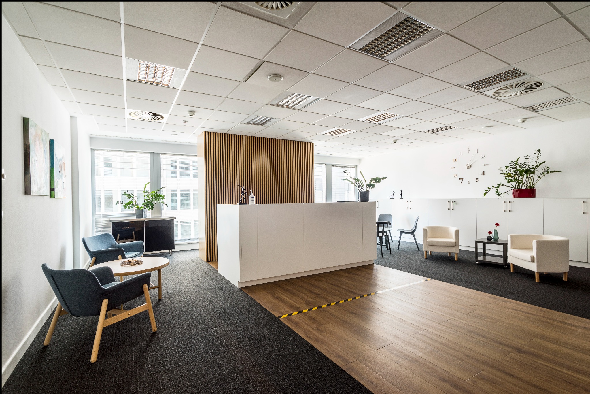 Jasne biuro z recepcją, strefą oczekiwania z niebieskimi fotelami i drewnianą podłogą. Minimalistyczny design, rośliny doniczkowe i zegar na ścianie.