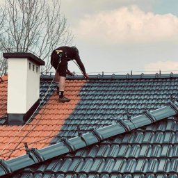 Top Service Adrian Wojniusz - Malowanie dachu z asekuracją linową. Widoczna część dachu pomalowana na grafitowo, reszta w kolorze ceglastym. Kominiarz przy kominie. Praca na wysokościach.