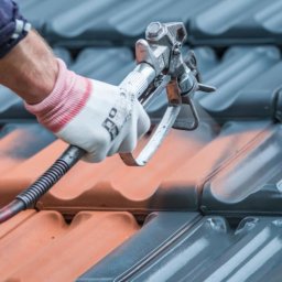 Top Service Adrian Wojniusz - Malowanie dachówki pistoletem natryskowym. Część dachu pomalowana na szaro, reszta w kolorze ceglastym. Ręka w rękawicy roboczej trzyma pistolet.