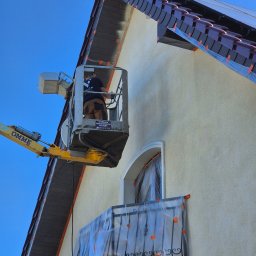 Top Service Adrian Wojniusz - Pracownik na podnośniku maluje elewację domu. Okna i balkon zabezpieczone folią ochronną. Widoczny fragment dachu z panelami słonecznymi. Słoneczny dzień.
