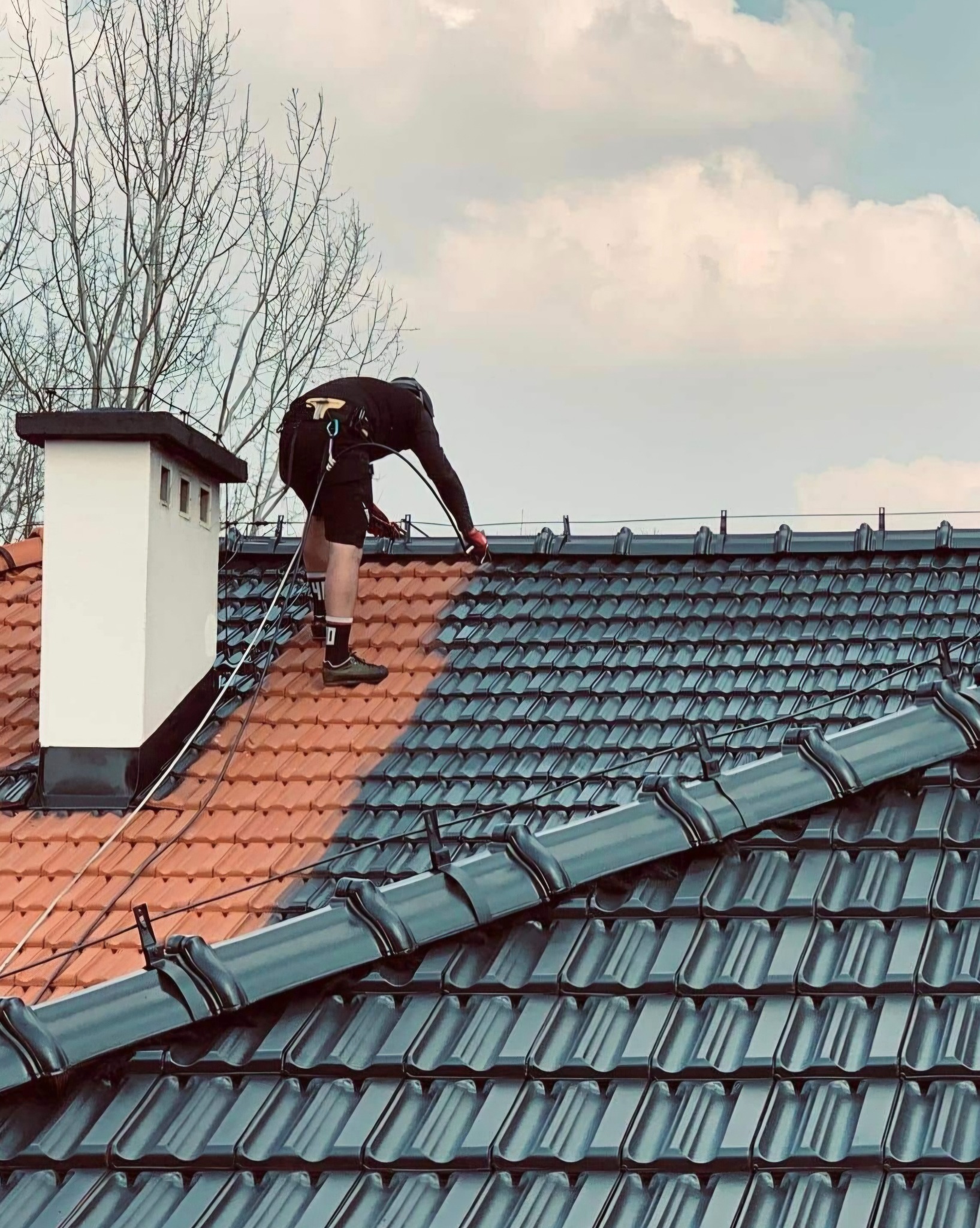 Malowanie dachu z asekuracją linową. Widoczna część dachu pomalowana na grafitowo, reszta w kolorze ceglastym. Kominiarz przy kominie. Praca na wysokościach.