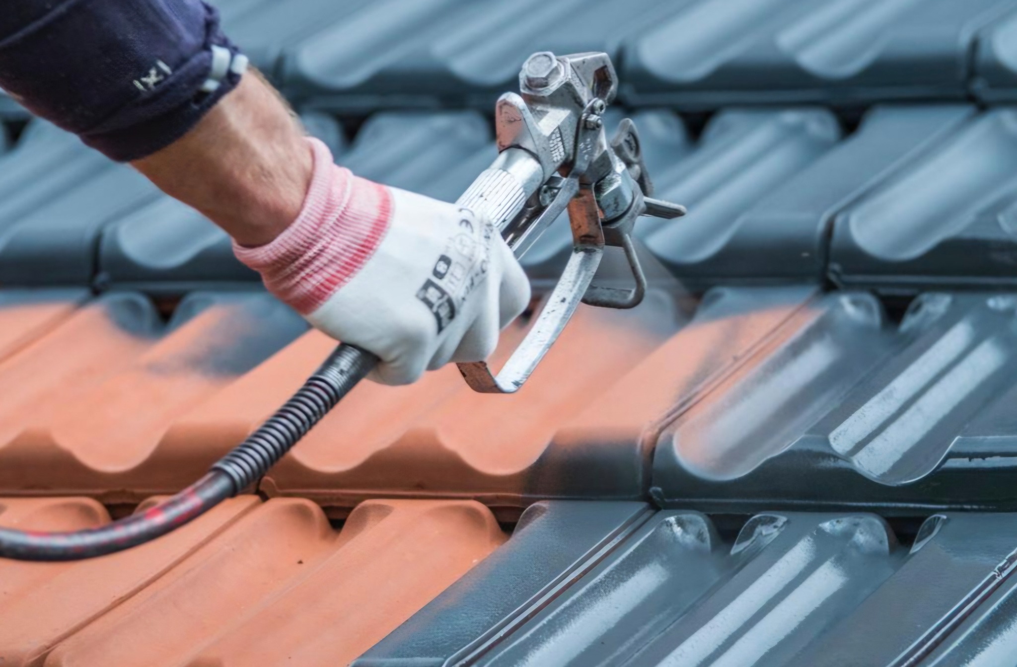 Malowanie dachówki pistoletem natryskowym. Część dachu pomalowana na szaro, reszta w kolorze ceglastym. Ręka w rękawicy roboczej trzyma pistolet.