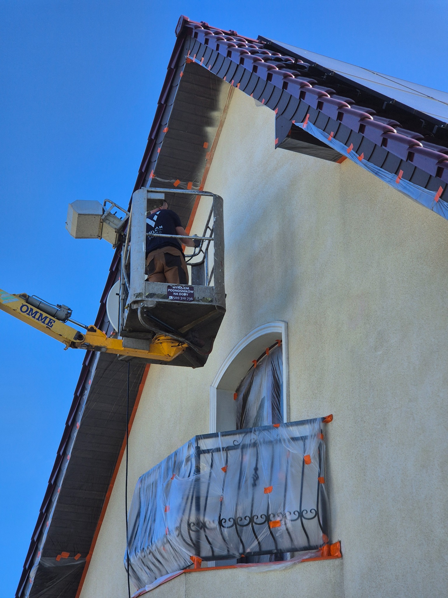 Pracownik na podnośniku maluje elewację domu. Okna i balkon zabezpieczone folią ochronną. Widoczny fragment dachu z panelami słonecznymi. Słoneczny dzień.