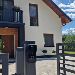 SMART EKO KAMIL WALASZKIEWICZ - Nowoczesny wideodomofon zamontowany na słupku bramy wjazdowej, na tle domu z balkonem i zadbanym podwórkiem. Widok z perspektywy otwartej bramy.