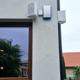 SMART EKO KAMIL WALASZKIEWICZ - Biała ściana budynku z zamontowanymi głośnikami zewnętrznymi i czujnikiem alarmowym z niebieską obwódką, widok z dołu, obok okno z odbiciem drzew.