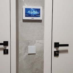 SMART EKO KAMIL WALASZKIEWICZ - Panel sterowania inteligentnego domu z dotykowym włącznikiem światła, zamontowany na ścianie wykończonej płytą imitującą marmur, pomiędzy białymi drzwiami.