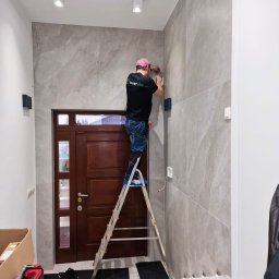 SMART EKO KAMIL WALASZKIEWICZ - Elektryk na drabinie montuje czujnik alarmowy na marmurowej ścianie w korytarzu. Widoczna skrzynka narzędziowa i wejście do domu.