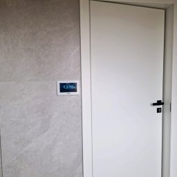 SMART EKO KAMIL WALASZKIEWICZ - Panel sterowania inteligentnego domu na szarej ścianie obok białych drzwi z czarną klamką. Widoczny czujnik i oświetlenie.