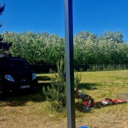 SMART EKO KAMIL WALASZKIEWICZ - Słup monitoringu z kamerami i niebieskim światłem ostrzegawczym na tle błękitnego nieba i zieleni. Widoczne narzędzia i samochód w tle.