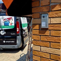 SMART EKO KAMIL WALASZKIEWICZ - Szary wideodomofon zamontowany na ceglanej ścianie domu, obok drabina i samochód firmy Smart-Eko z reklamą systemów alarmowych i wideodomofonów.