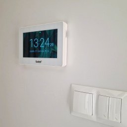 SMART EKO KAMIL WALASZKIEWICZ - Biały panel dotykowy Safel z wyświetlaną godziną i datą, zamontowany na ścianie obok włącznika światła i dekoracyjnych paneli ściennych.