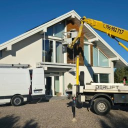 SMART EKO KAMIL WALASZKIEWICZ - Żółty podnośnik koszowy PERLIFT przy nowoczesnym domu z dużymi oknami. Biały bus z otwartymi drzwiami obok. Antena satelitarna na ścianie. Słoneczny dzień.