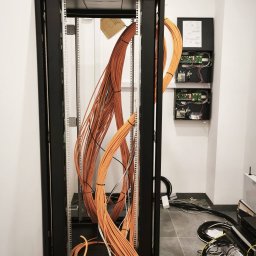 SMART EKO KAMIL WALASZKIEWICZ - Szafa rack z pomarańczowymi kablami w trakcie instalacji. Na ścianie skrzynka z elektroniką. Widoczne przewody i elementy montażowe. Szara podłoga.