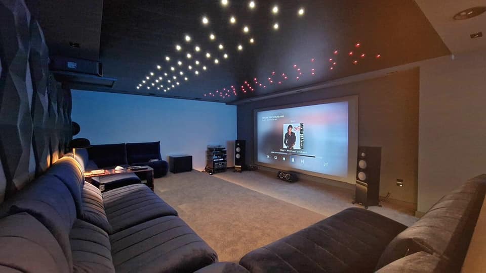 Kino domowe z projektorem, nagłośnieniem i oświetleniem punktowym na suficie. Na ekranie odtwarzany jest utwór Michaela Jacksona. Wnętrze w ciemnych barwach.