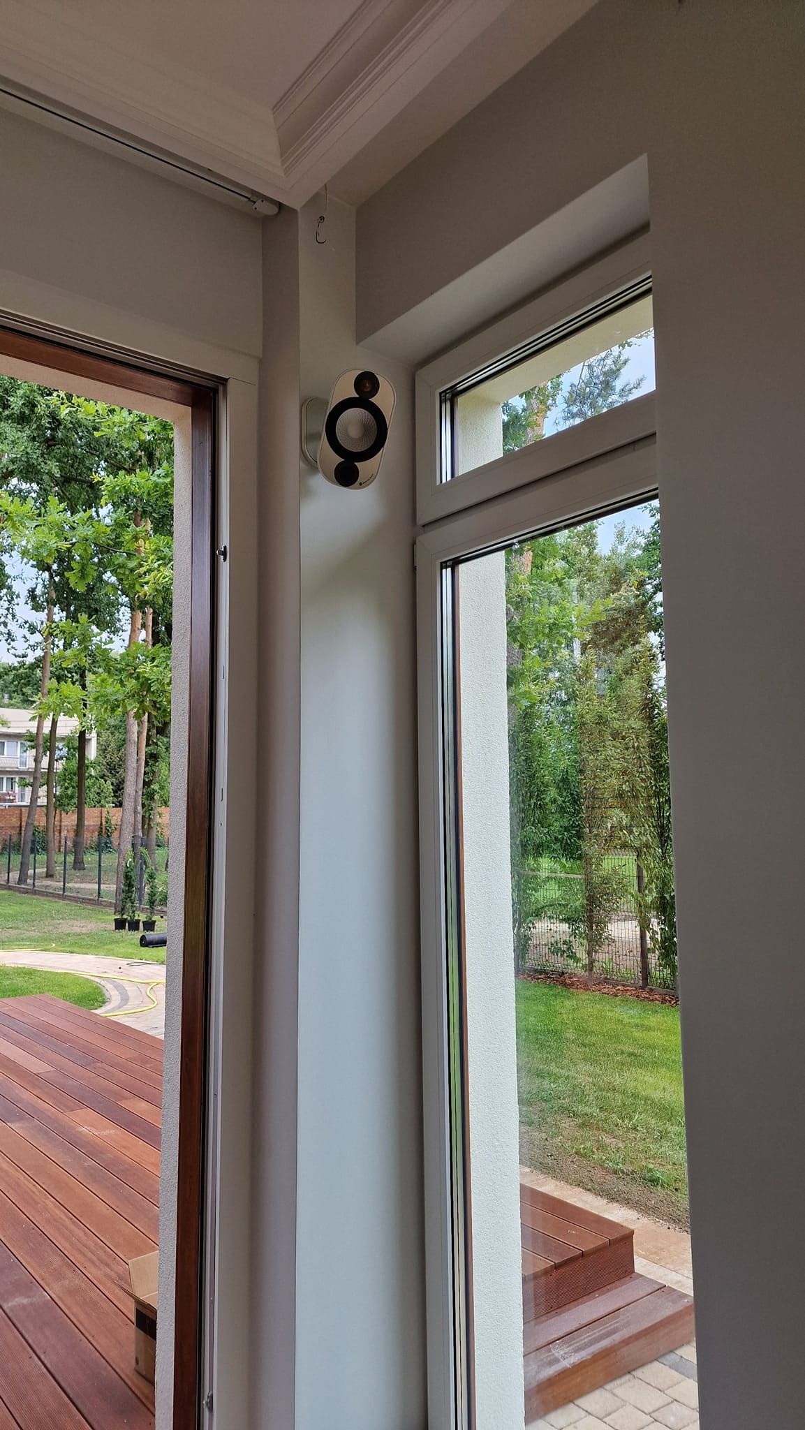 Kamera monitoringu zamontowana w rogu pokoju, widok na zewnątrz przez okno i drzwi balkonowe. Biała ściana, drewniany taras na zewnątrz.