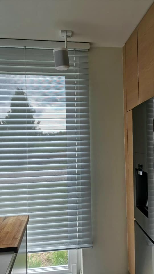 Szary głośnik systemu inteligentnego domu zamontowany na suficie, widoczny na tle okna z żaluzjami i fragmentu zabudowy kuchennej.