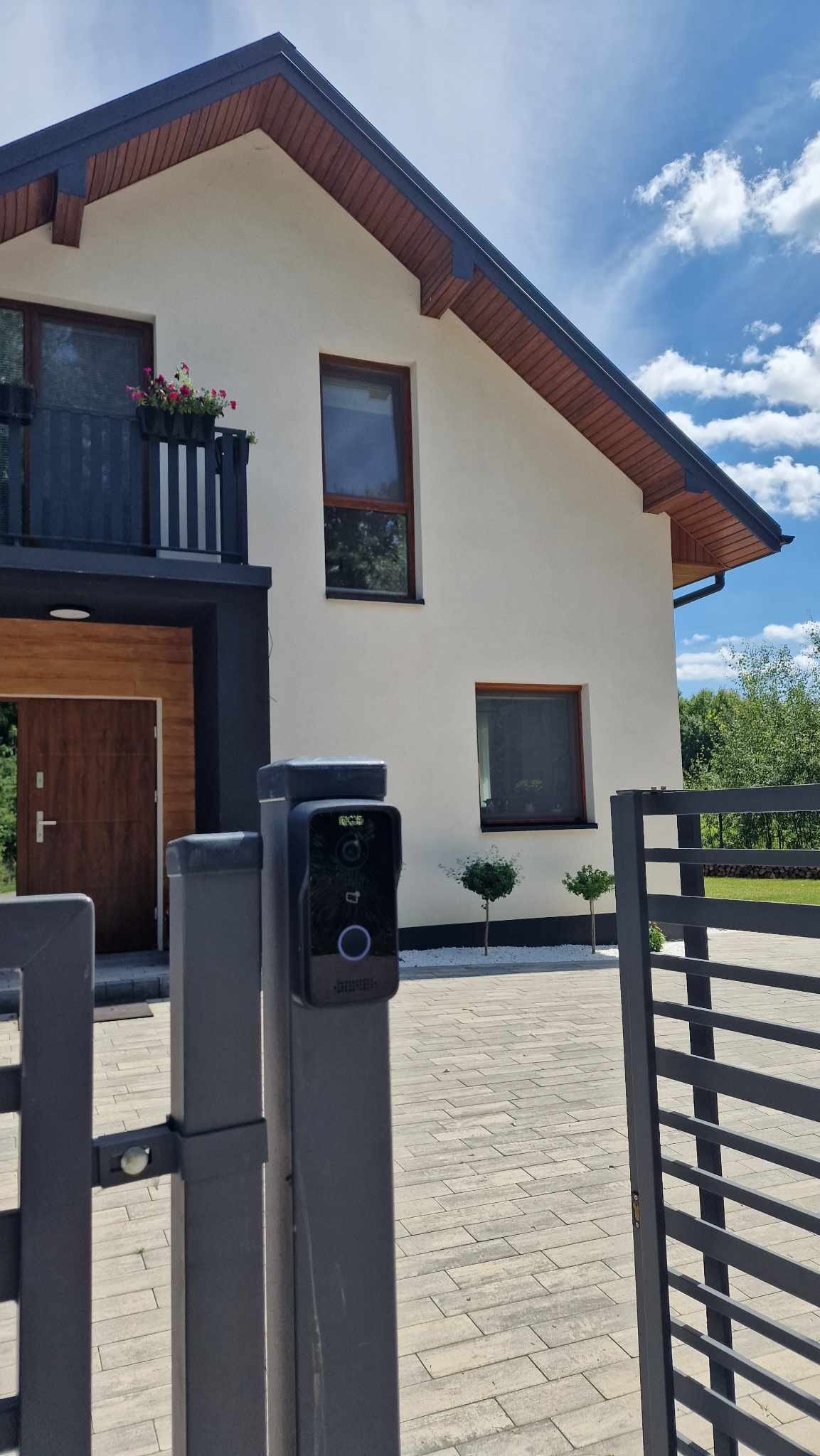 Nowoczesny wideodomofon zamontowany na słupku bramy wjazdowej, na tle domu z balkonem i zadbanym podwórkiem. Widok z perspektywy otwartej bramy.