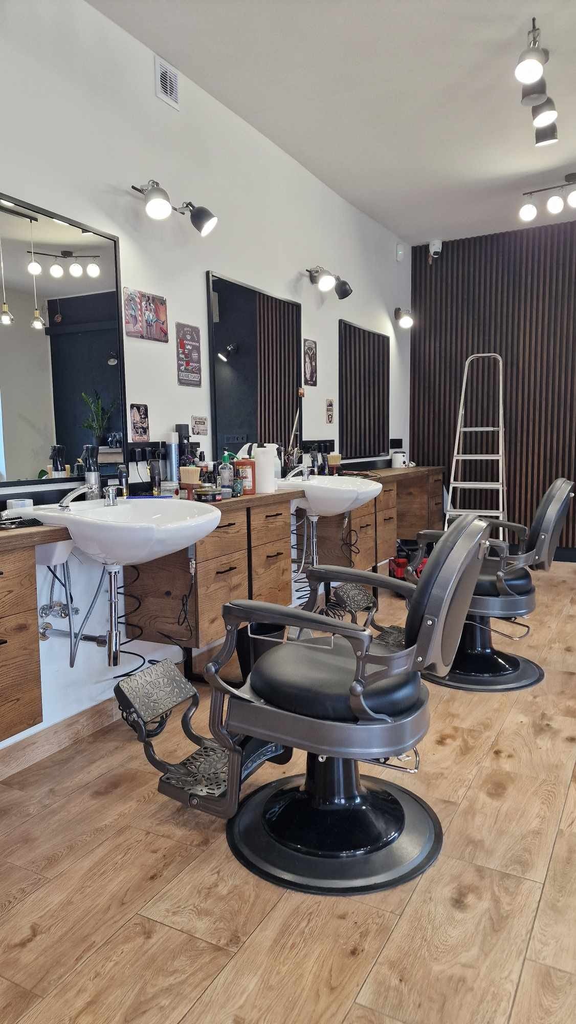 Nowoczesne wnętrze salonu fryzjerskiego: dwa fotele barberskie, umywalki z drewnianymi szafkami, lustra i oświetlenie punktowe. Na ścianie dekoracyjne panele.