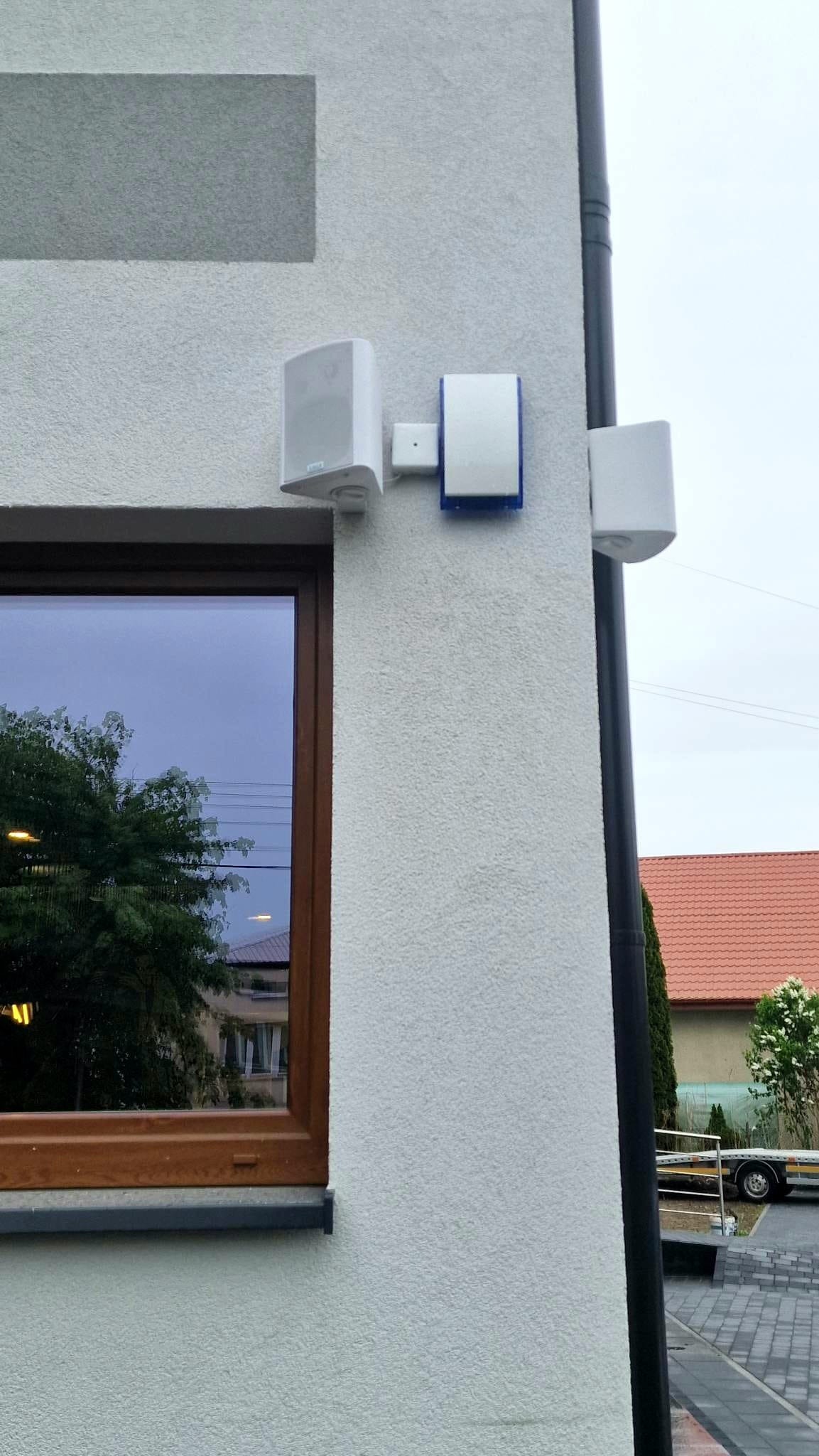 Biała ściana budynku z zamontowanymi głośnikami zewnętrznymi i czujnikiem alarmowym z niebieską obwódką, widok z dołu, obok okno z odbiciem drzew.