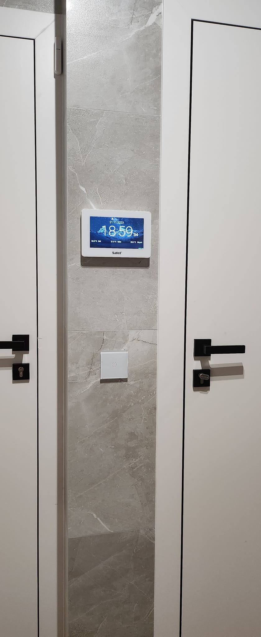 Panel sterowania inteligentnego domu z dotykowym włącznikiem światła, zamontowany na ścianie wykończonej płytą imitującą marmur, pomiędzy białymi drzwiami.